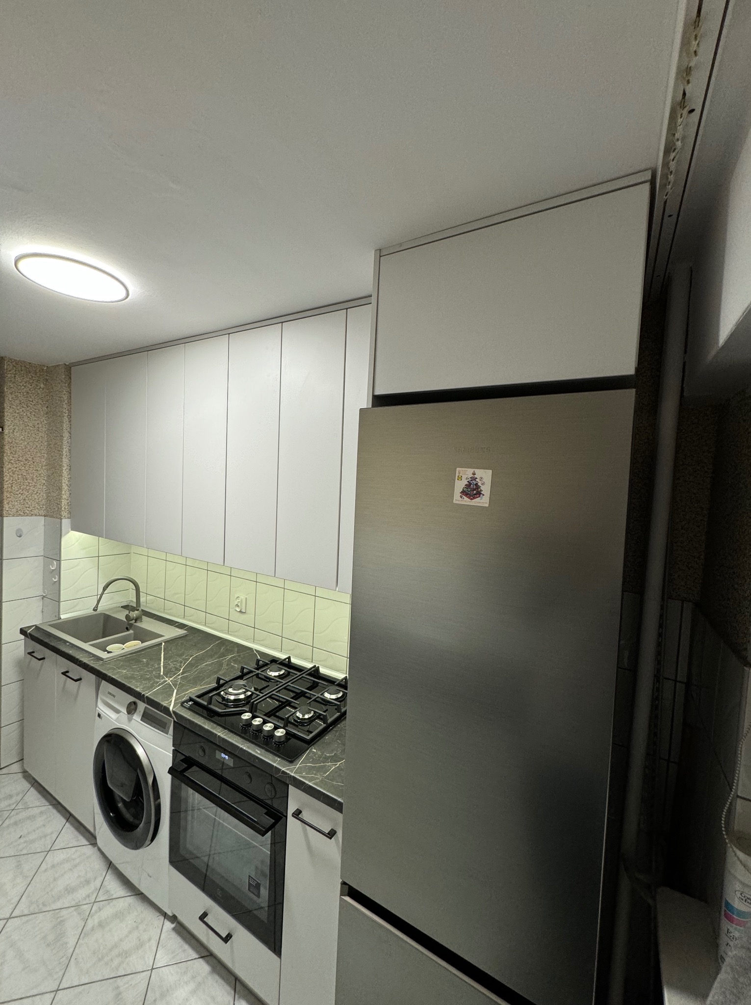 Wnętrze małej kuchni z szarymi szafkami, czarnym blatem, pralką obok zlewu i lodówką z magnesem na drzwiach.