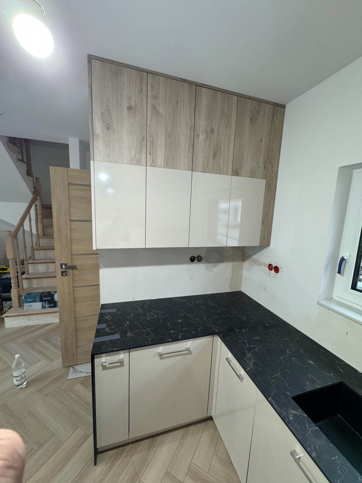 Fragment kuchni w trakcie montażu: białe szafki dolne i górne z drewnopodobnymi frontami, czarny blat z marmurkowym wzorem, widoczne instalacje elektryczne i hydrauliczne w ścianie, podłoga w jodełkę.