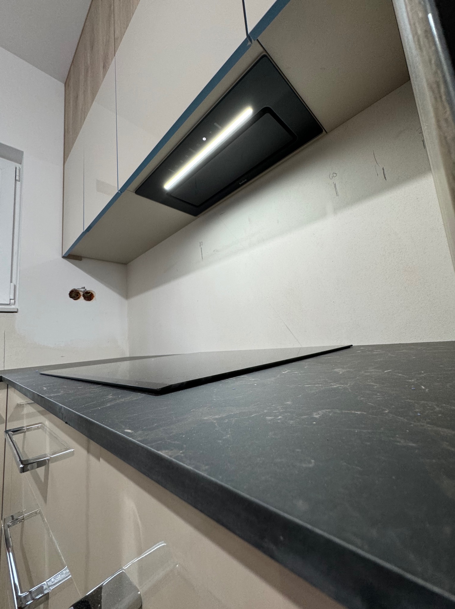 Fragment kuchni w trakcie montażu: widoczny blat z czarnego kamienia, płyta indukcyjna, szafki w jasnym odcieniu beżu z chromowanymi uchwytami oraz okap podszafkowy z oświetleniem LED. Na ścianie...
