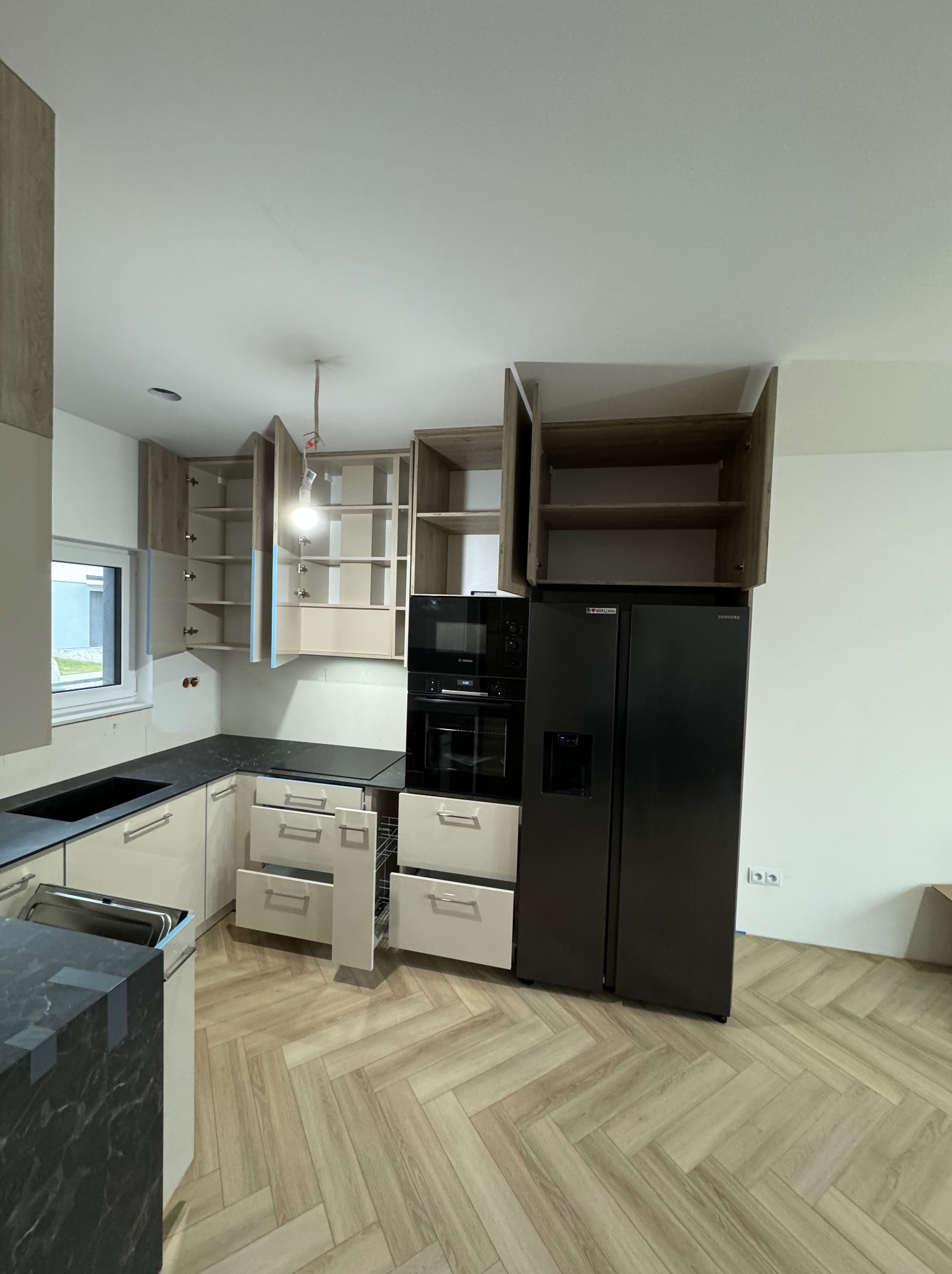 Montaż kuchni: beżowe szafki z czarnym blatem, otwarte górne szafki z półkami, czarny piekarnik i lodówka, parkiet w jodełkę, widoczne kable oświetleniowe.