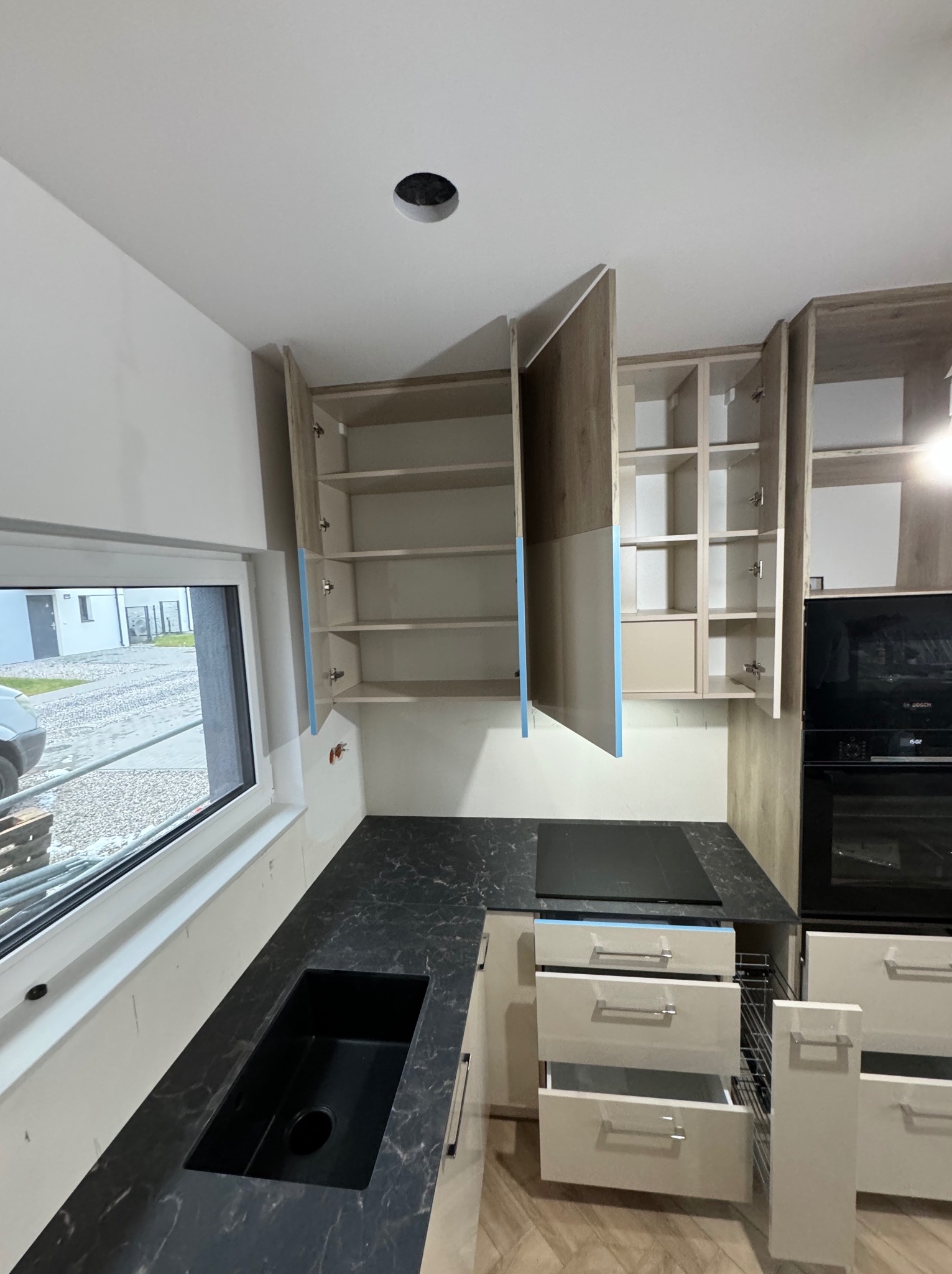 Nowoczesna kuchnia w trakcie montażu: otwarte szafki wiszące z półkami, czarny blat z wbudowanym zlewem i płytą indukcyjną, wysuwane szuflady z metalowymi koszami, widoczne zabezpieczenia...