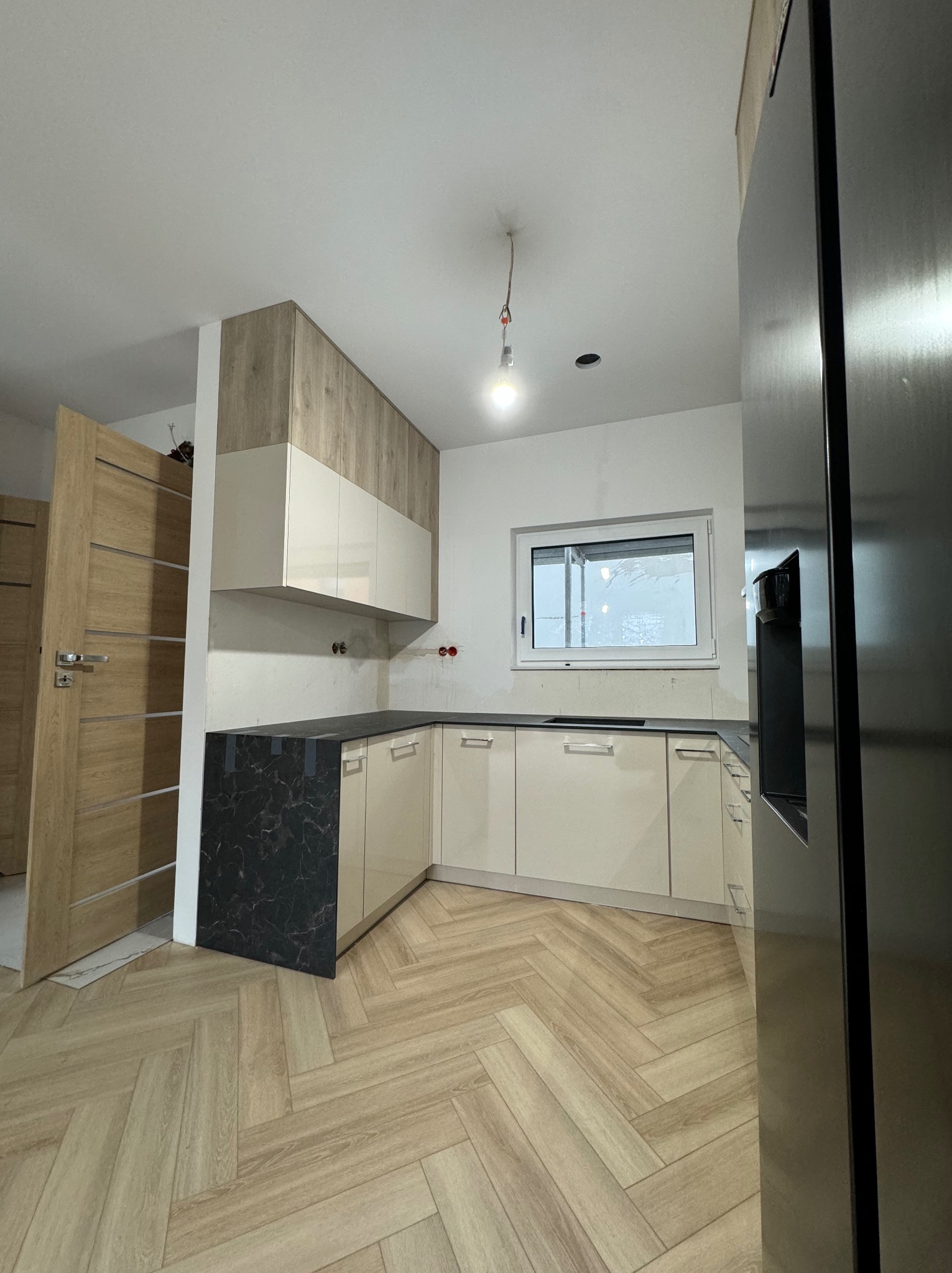 Kremowa kuchnia na wymiar z czarnym blatem i drewnianymi akcentami, widok na podłogę ułożoną w jodełkę, częściowo widoczna lodówka.