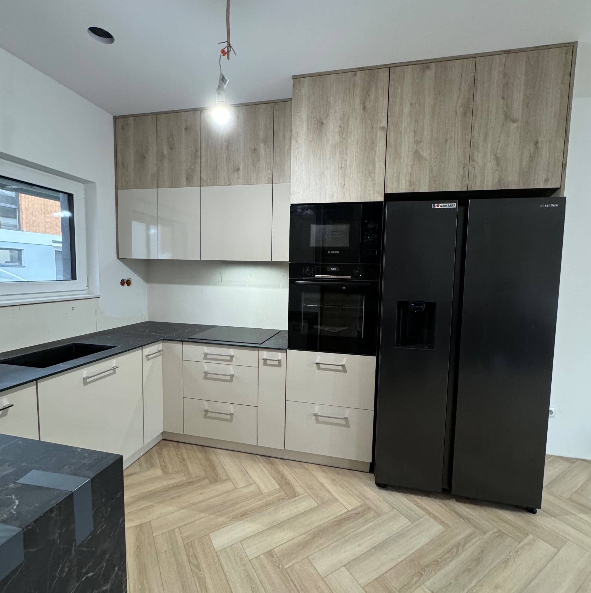 Nowoczesna kuchnia w trakcie montażu: biało-drewniane szafki, czarny blat i sprzęt AGD, podłoga w jodełkę, widoczne niedokończone prace wykończeniowe.