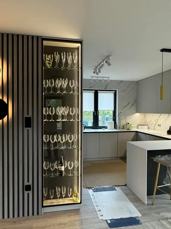 Nowoczesna kuchnia z szarą zabudową i marmurowym blatem, wysoka przeszklona szafka z podświetleniem wypełniona kieliszkami, widoczne elementy wykończenia ścian w pionowe pasy.