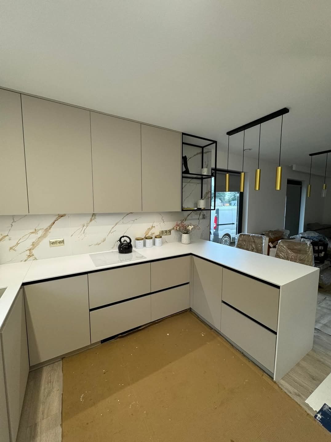 Jasna, narożna zabudowa kuchenna z białym blatem i marmurowym backsplashiem, czarne, minimalistyczne uchwyty, widoczne złote lampy wiszące i fragment transportowego kartonu na podłodze.