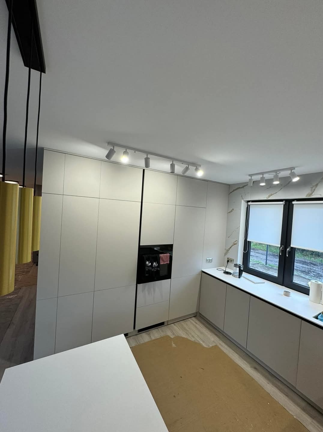 Nowoczesna kuchnia z zabudową w kolorze jasnej szarości, oświetleniem punktowym i marmurowym akcentem na ścianie, widok na okno z roletami.