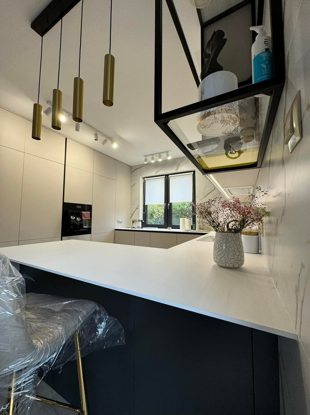 Nowoczesna kuchnia w Warszawie z białym blatem, czarnymi akcentami i designerskim oświetleniem. Krzesło barowe owinięte folią, marmurowe wykończenie ściany oraz dekoracyjne kwiaty w wazonie.