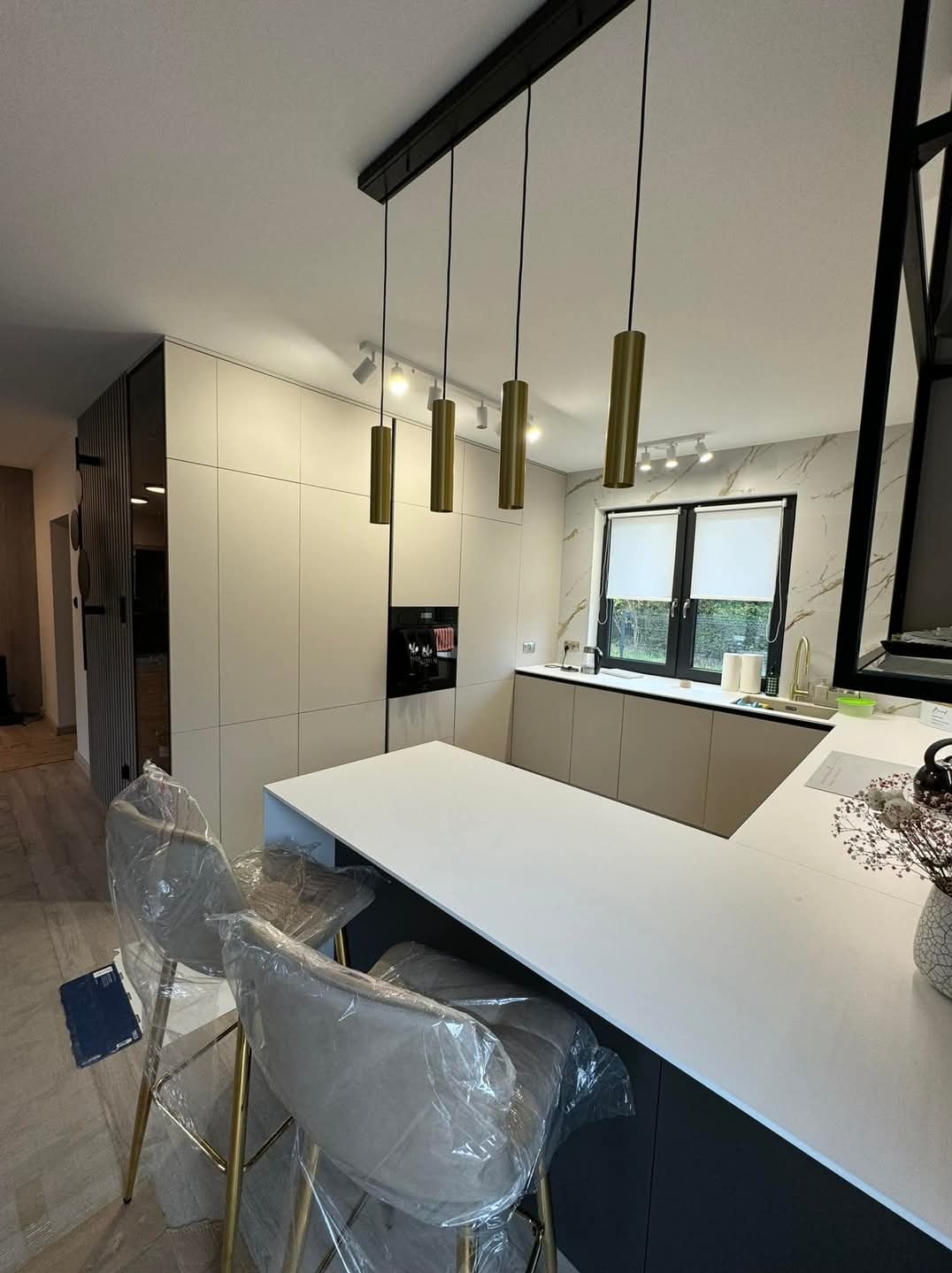 Nowoczesna kuchnia z beżowymi szafkami, białym blatem wyspy i złotymi lampami wiszącymi. Krzesła barowe z złotymi nogami zabezpieczone folią ochronną.