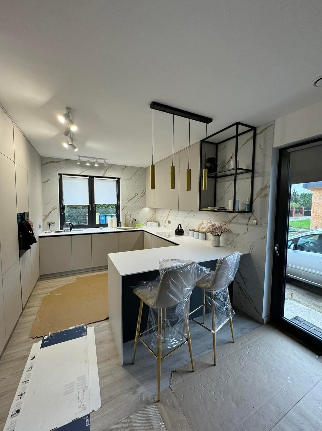 Nowoczesna kuchnia z wyspą, wykończona marmurowymi płytami, z wiszącymi złotymi lampami i czarnym regałem; krzesła barowe zabezpieczone folią, podłoga częściowo zakryta kartonem.
