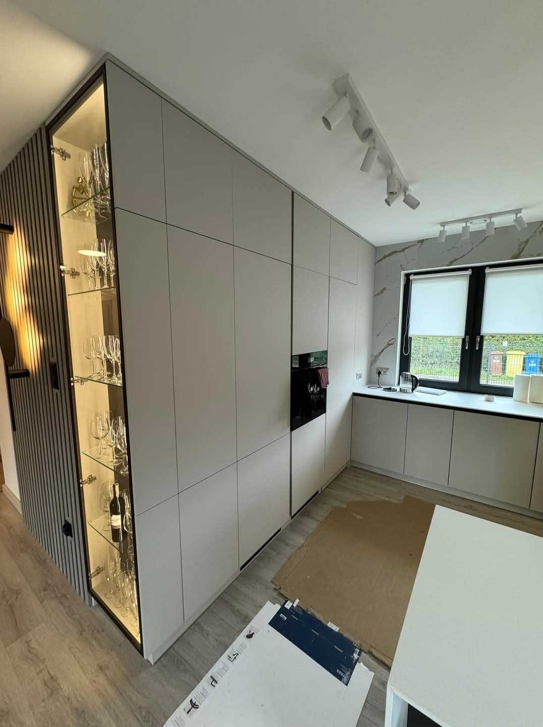 Nowoczesna kuchnia z zabudową w odcieniach szarości, podświetlana witryna z kieliszkami, minimalistyczne oświetlenie sufitowe i marmurowe wykończenie ściany przy oknie.