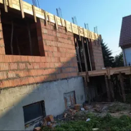 Wznoszenie muru z czerwonej cegły porotherm na fundamencie z betonu komórkowego, widoczne drewniane szalunki stropu i zbrojenie, w tle budynek mieszkalny i żółta koparka.