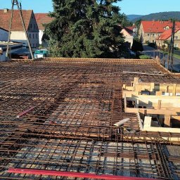 Nikolas słomski - Zbrojenie stropu budynku w trakcie budowy z widocznymi szalunkami drewnianymi i elementami konstrukcyjnymi na tle zabudowy mieszkalnej.