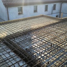 Nikolas słomski - Zbrojenie stropu przygotowane do zalania betonem, widoczne szalunki i wystające pręty, w tle budynek mieszkalny.