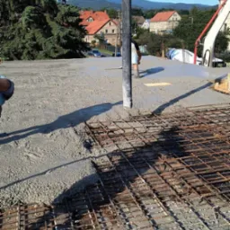 Wylewanie betonu na zbrojenie fundamentu domu jednorodzinnego, widoczna pompa do betonu, domy w tle.