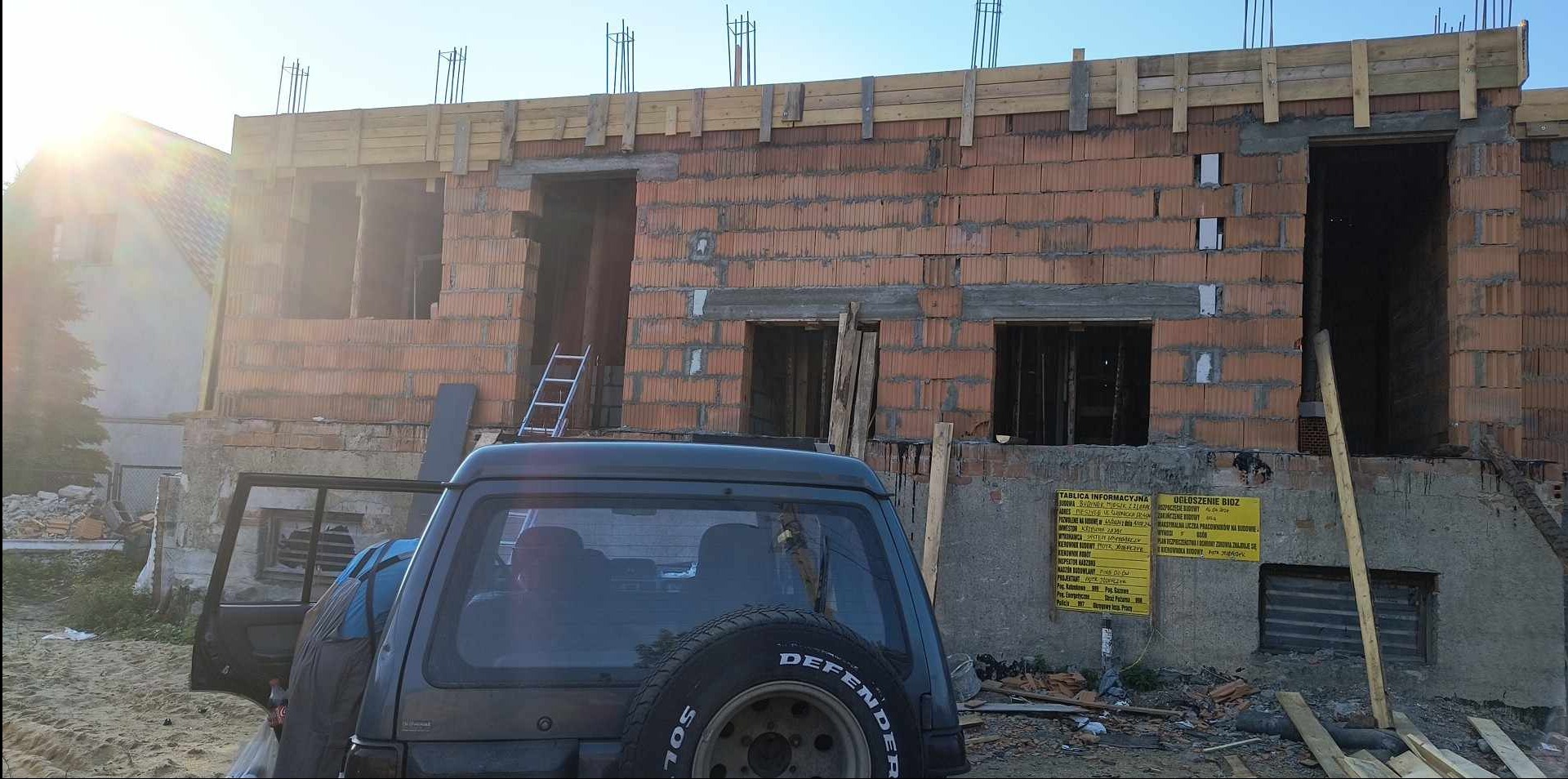 Wznoszenie murów z pustaków ceramicznych na budowie domu jednorodzinnego, widoczne zbrojenia i drewniane szalunki, w tle zaparkowany samochód terenowy z otwartymi drzwiami.
