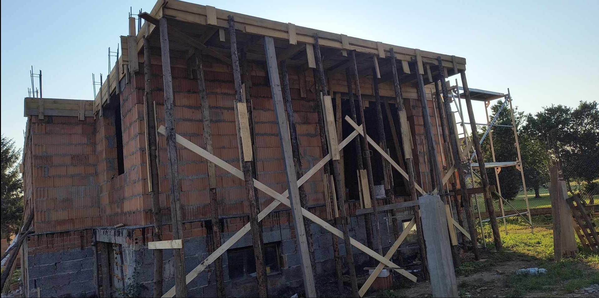 Surowa konstrukcja budynku z czerwonej cegły, wzmocniona drewnianymi stemplami i szalunkami, w tle drzewa i niebieskie niebo.
