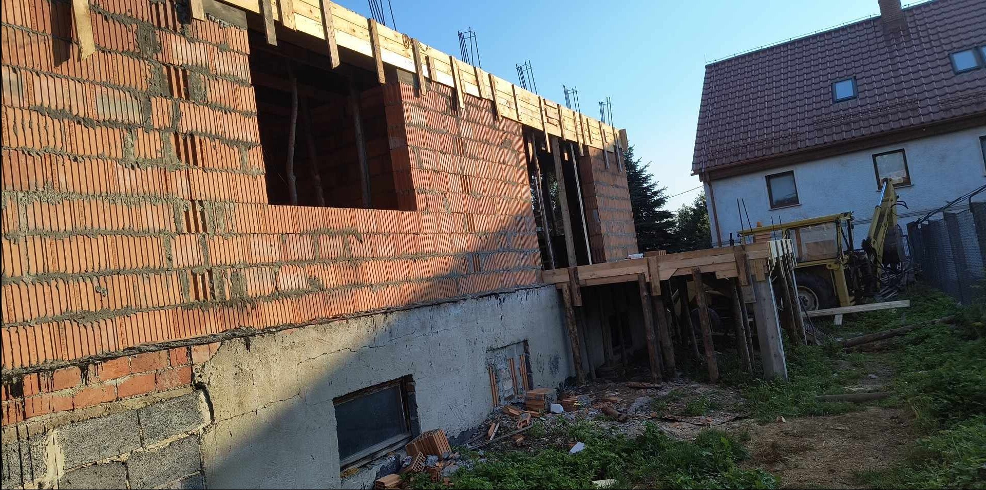 Wznoszenie muru z czerwonej cegły porotherm na fundamencie z betonu komórkowego, widoczne drewniane szalunki stropu i zbrojenie, w tle budynek mieszkalny i żółta koparka.