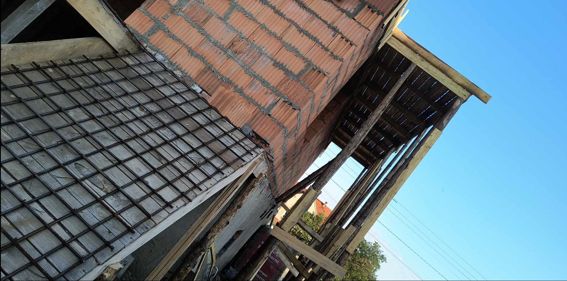 Wznoszenie muru z czerwonej cegły ceramicznej, widoczna konstrukcja drewniana szalunku oraz zbrojenie betonu na budowie domu jednorodzinnego, niebo w tle.