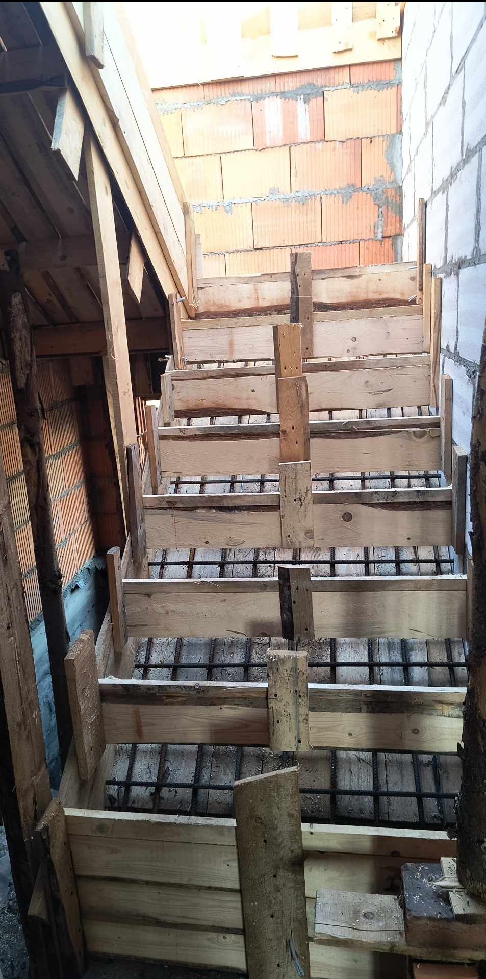 Formowanie drewniane pod schody betonowe, widoczne zbrojenie stalowe, w tle ściana z czerwonej cegły dziurawki i ściana z białych bloczków gazobetonowych.