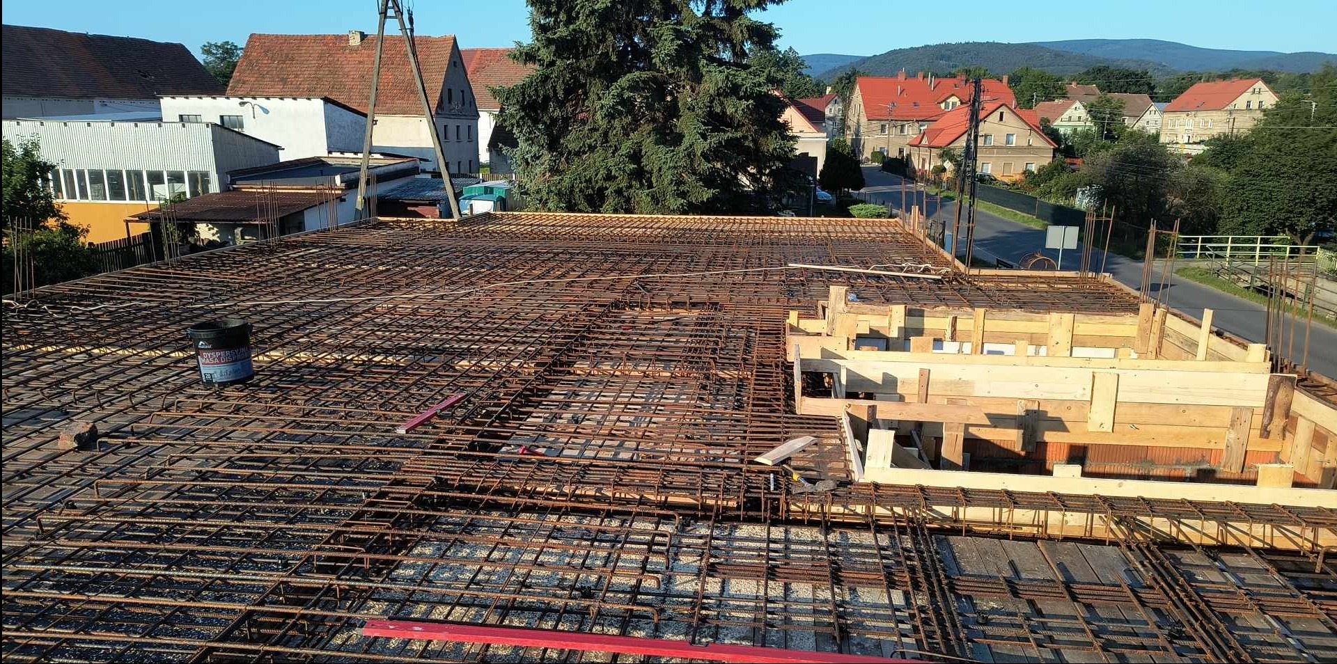 Zbrojenie stropu budynku w trakcie budowy z widocznymi szalunkami drewnianymi i elementami konstrukcyjnymi na tle zabudowy mieszkalnej.