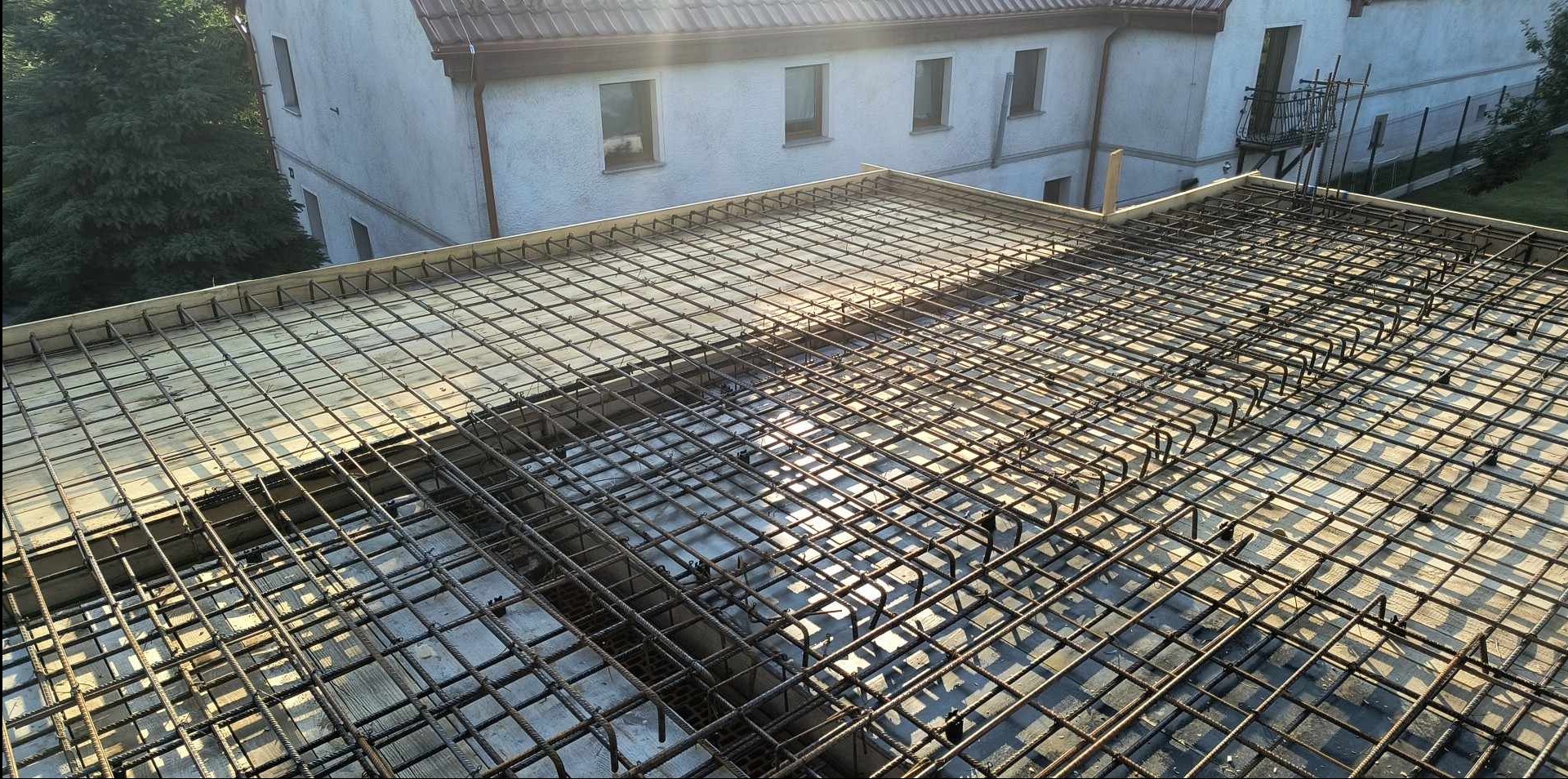 Zbrojenie stropu przygotowane do zalania betonem, widoczne szalunki i wystające pręty, w tle budynek mieszkalny.