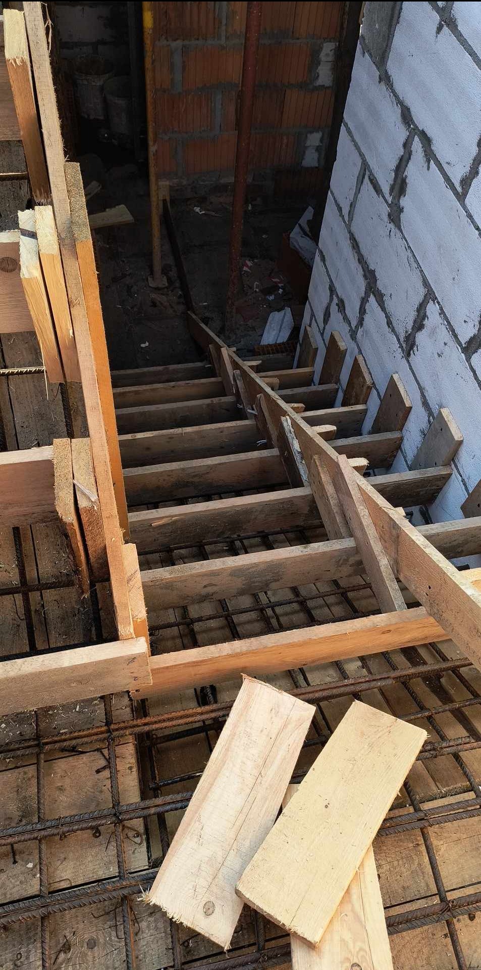 Szalunek schodów w trakcie budowy, widoczne zbrojenie i fragment murowanej ściany z bloczków. Drewniane elementy konstrukcji tymczasowej.