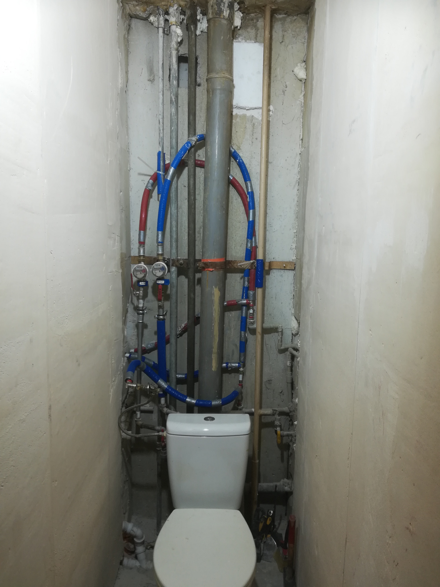 Toaleta z widocznymi rurami wodnymi w trakcie montażu w surowej łazience przed wykończeniem, z odsłoniętymi przewodami i miernikami.