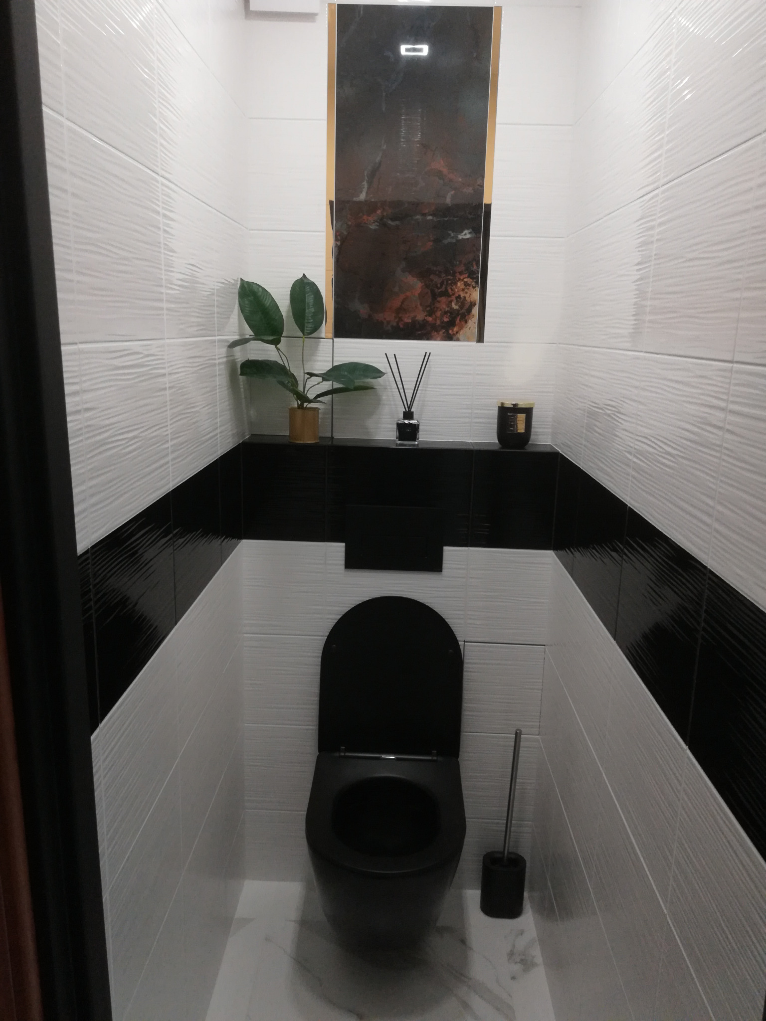 Nowoczesna toaleta z białymi, falistymi płytkami ściennymi, czarnym pasem dekoracyjnym, czarną armaturą i marmurową podłogą, uzupełniona o roślinę doniczkową i akcesoria zapachowe na półce.