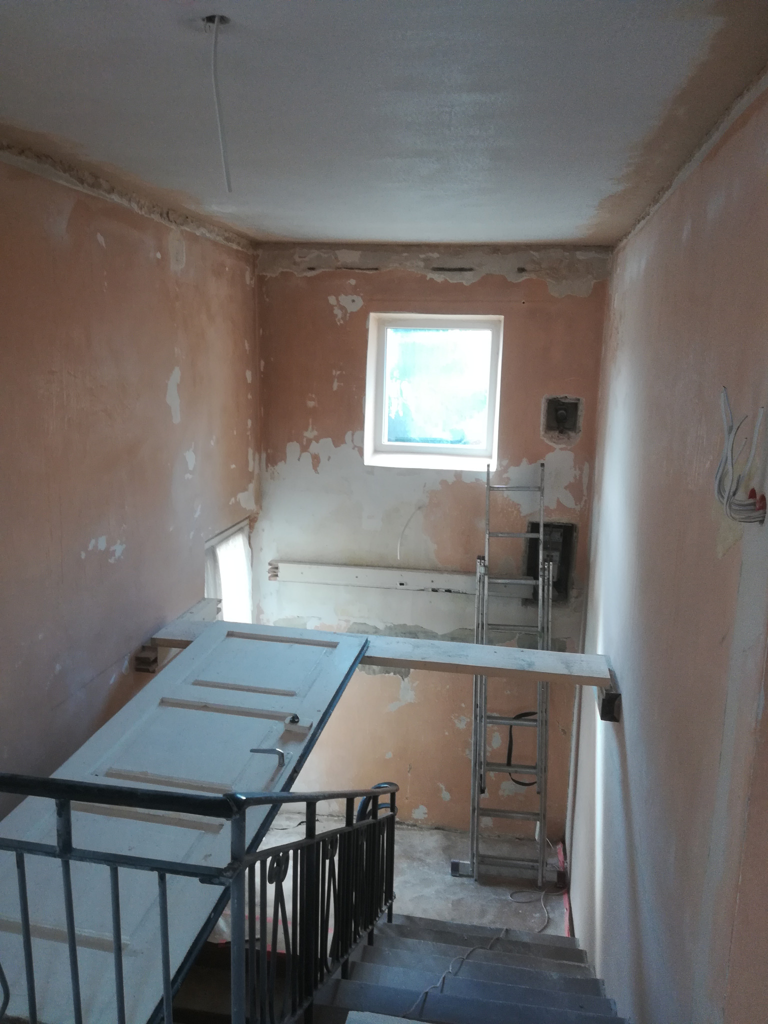 Wnętrze w trakcie remontu: ściany z resztkami starej farby, drabina, prowizoryczna kładka z desek i drzwi na schodach, instalacja elektryczna wystająca ze ściany.