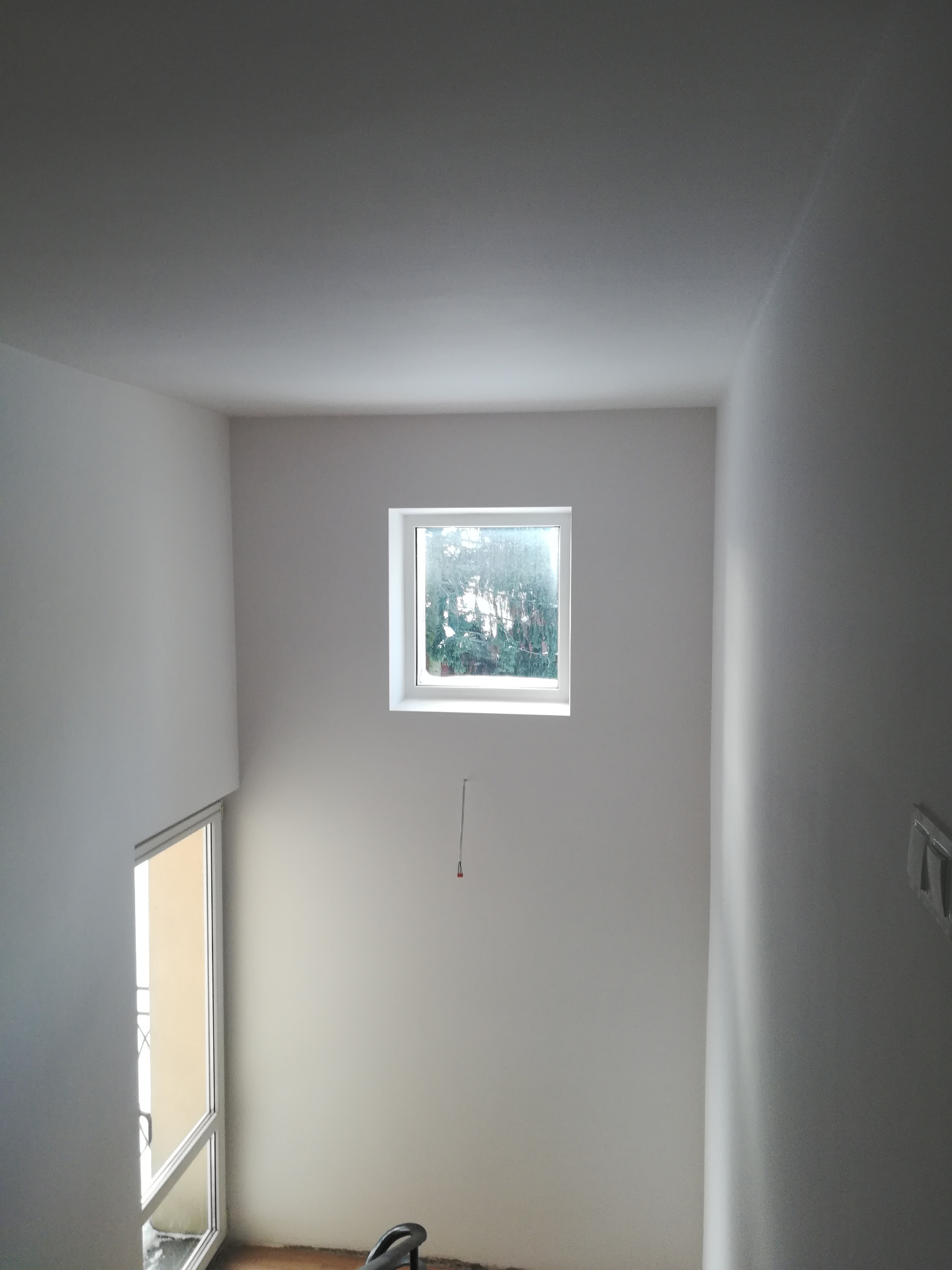 Wnętrze klatki schodowej po świeżym malowaniu na biało, widoczne okno z widokiem na zieleń, fragment drzwi balkonowych i nieduży element drabiny malarskiej na podłodze.