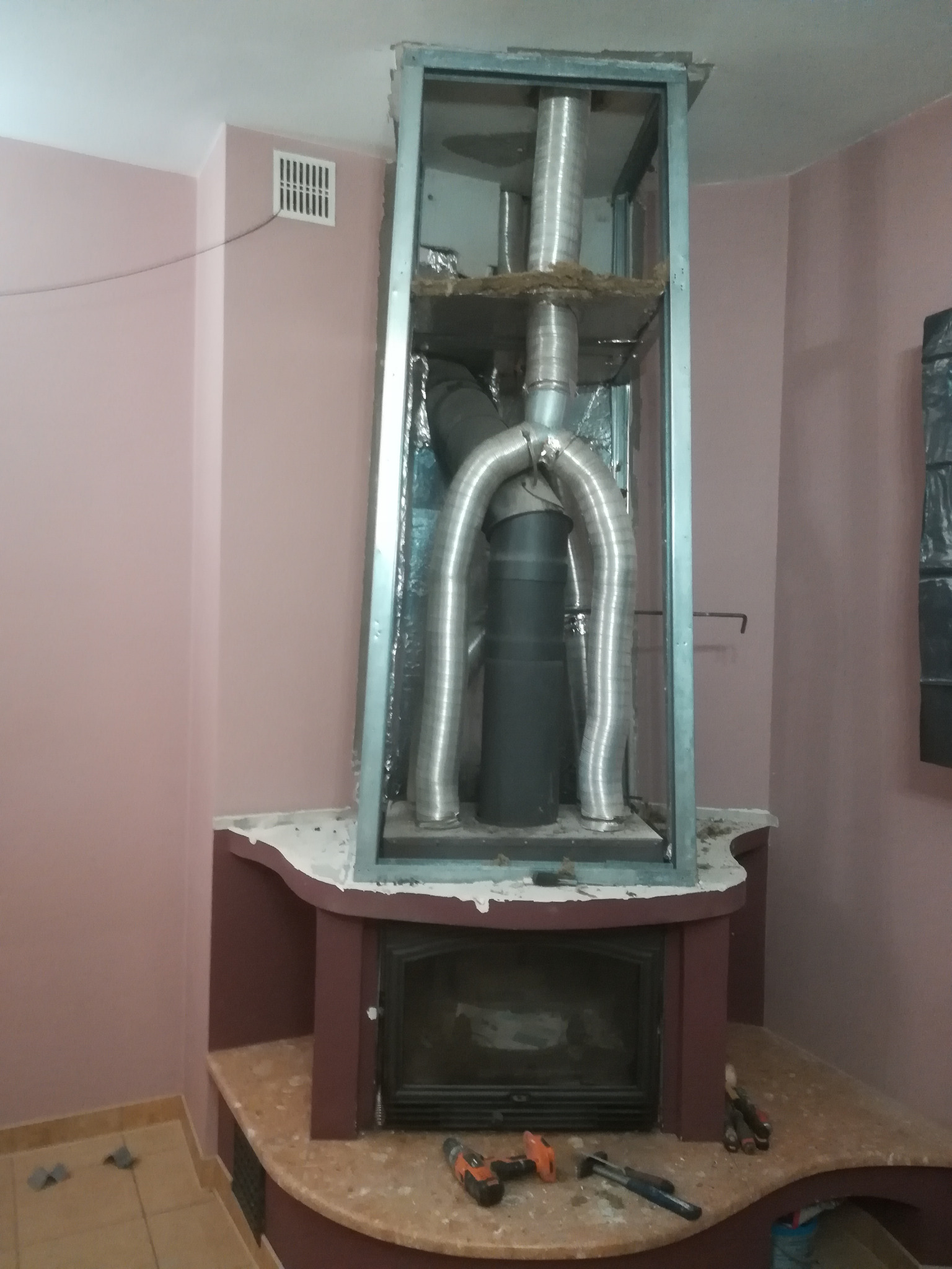 Montaż wkładu kominkowego z widocznymi rurami spalinowymi w metalowej ramie, narzędzia leżące na obudowie kominka, ściany w odcieniach różu.