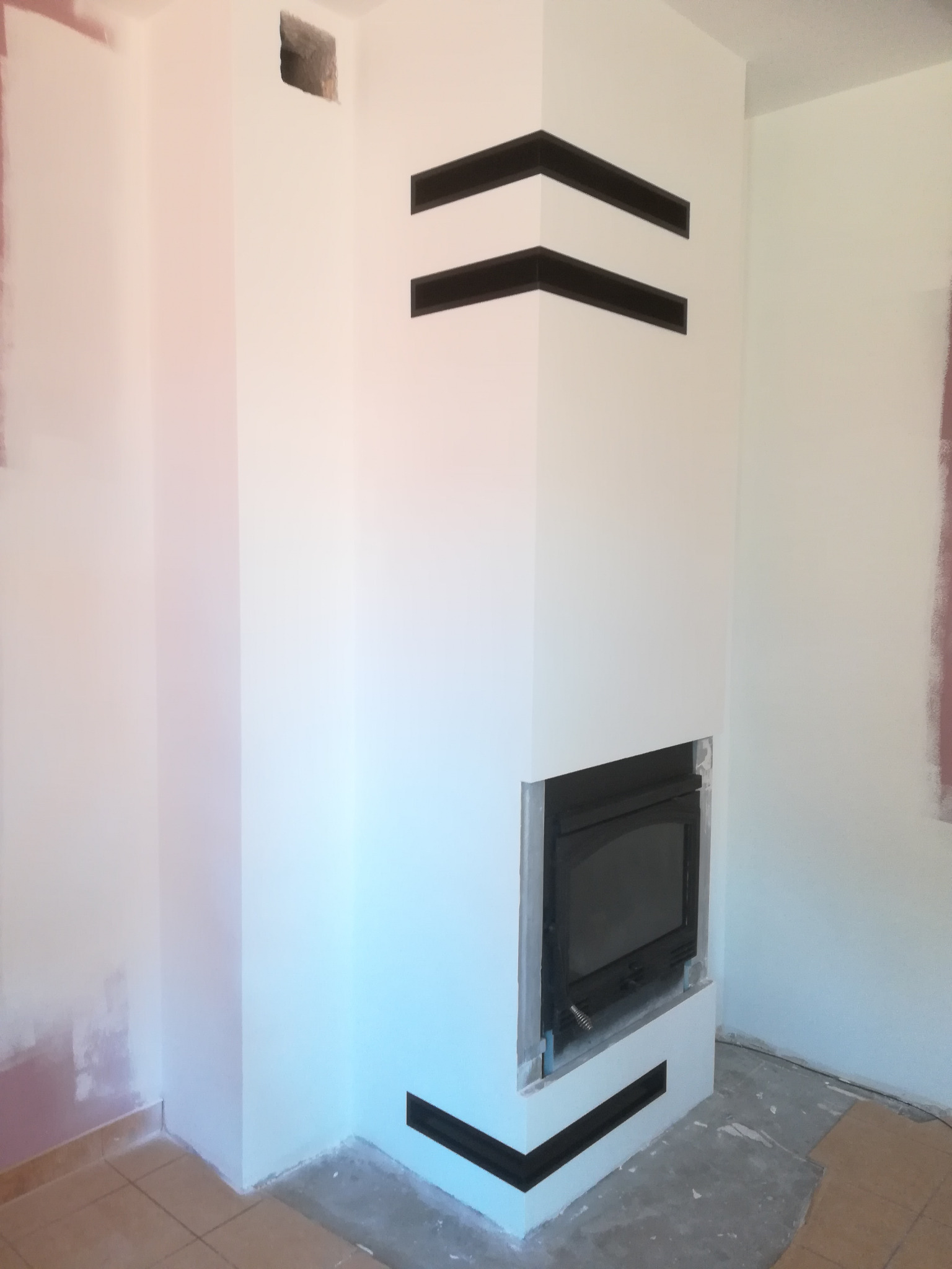 Biały kominek z czarnymi, poziomymi akcentami dekoracyjnymi, w trakcie wykończenia, z widocznymi śladami szpachlowania na ścianach i brakiem listew przypodłogowych.