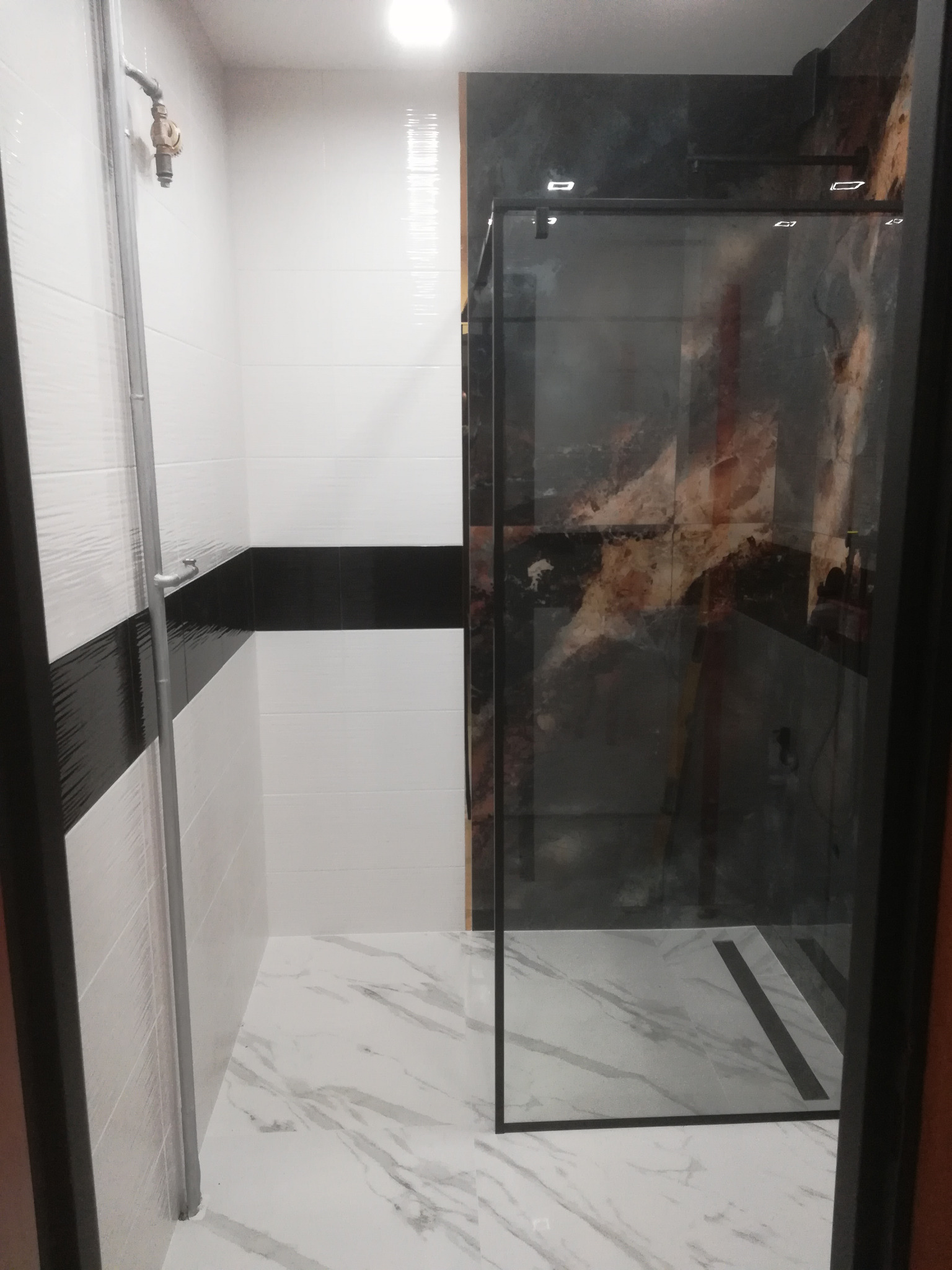 Nowoczesna łazienka z kabiną prysznicową typu walk-in, biało-czarne płytki na ścianach z akcentem imitacji marmuru, marmurowa podłoga oraz widoczna bateria prysznicowa.