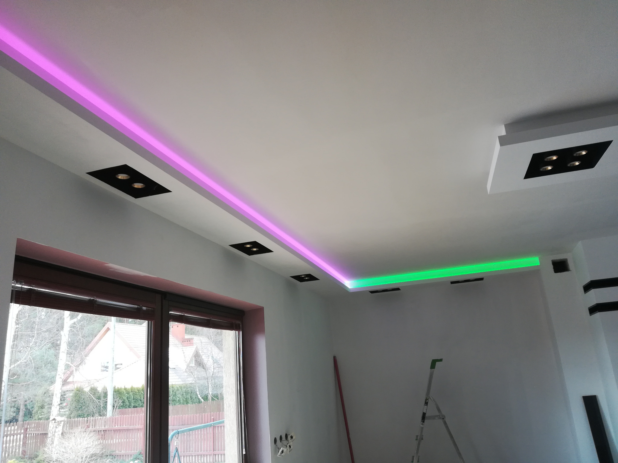 Nowoczesny sufit podwieszany z oświetleniem LED w kolorach różowym i zielonym, czarne oprawy oświetleniowe, widoczne niedokończone ściany i drabina.