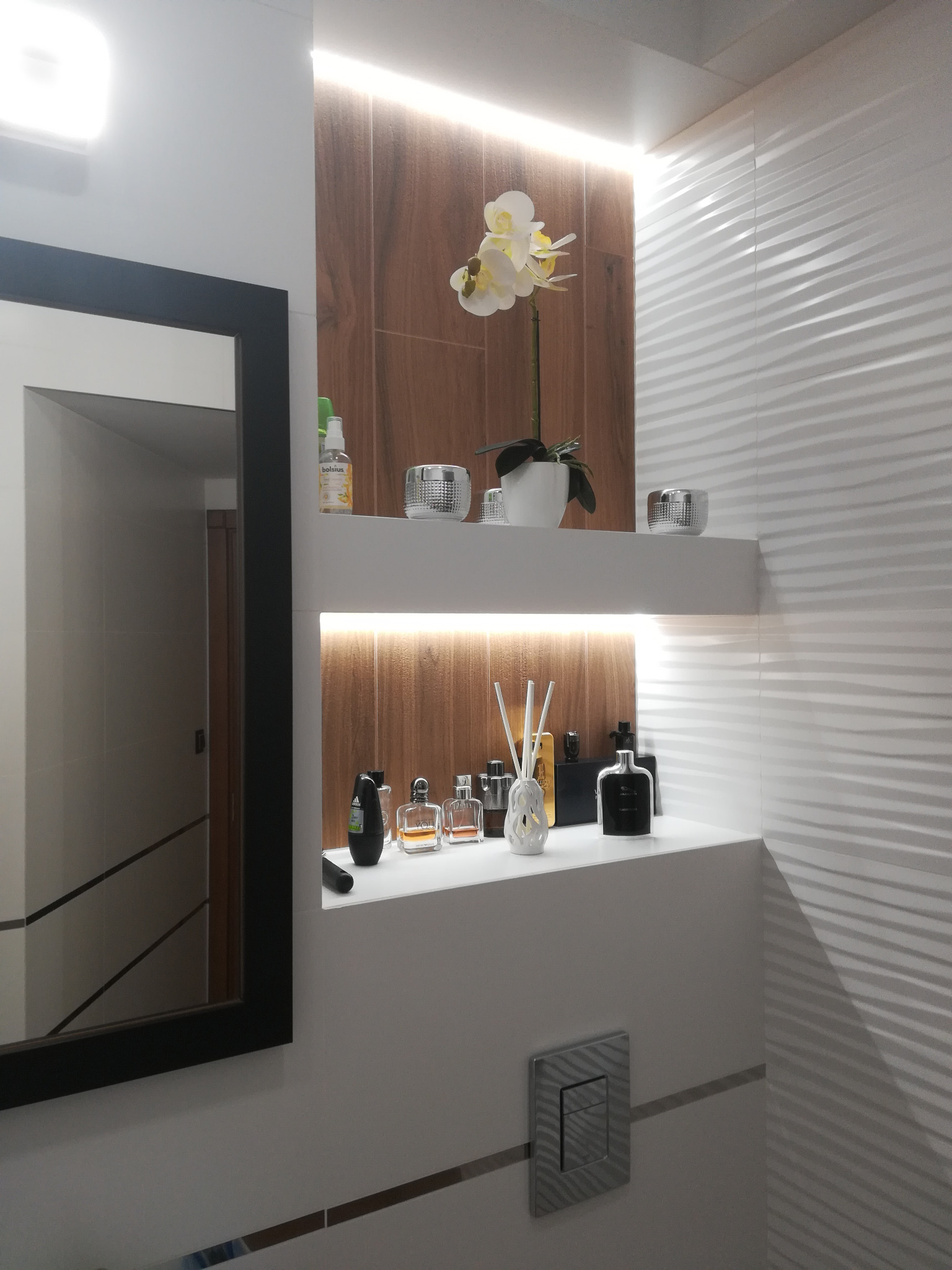 Nowoczesna łazienka z półkami wnękowymi podświetlonymi LED, dekoracyjną orchideą i flakonami perfum, białe płytki faliste na ścianie, czarna rama lustra.