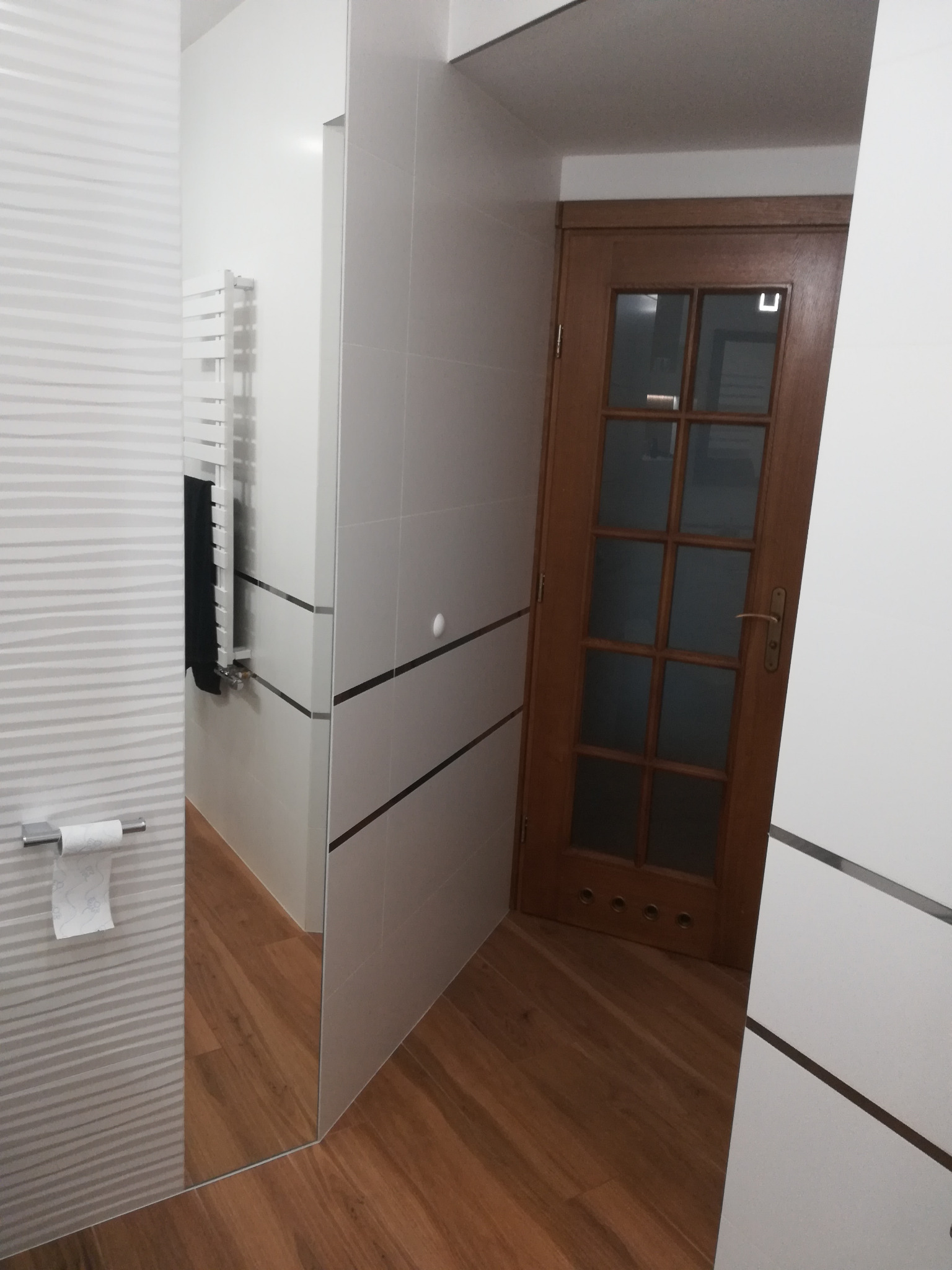 Nowoczesna łazienka z białymi płytkami o falistej strukturze, drewnianą podłogą i drzwiami z przeszkleniami, widoczny grzejnik drabinkowy i lustro.