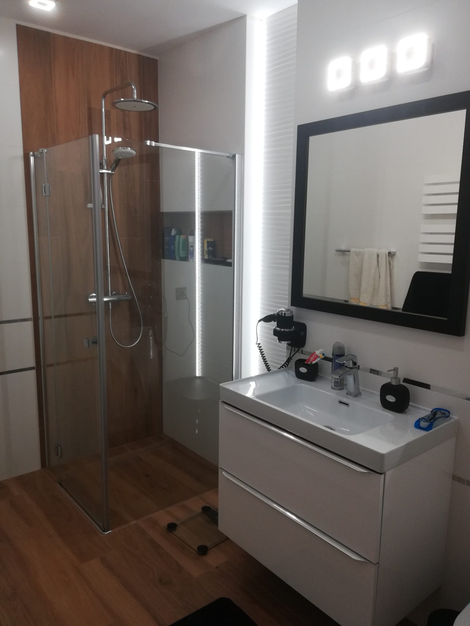 Nowoczesna łazienka z kabiną prysznicową typu walk-in z deszczownicą, umywalką z szafką w kolorze białym, lustrem w czarnej ramie i trzema kwadratowymi lampami LED na ścianie, drewnopodobne płytki...