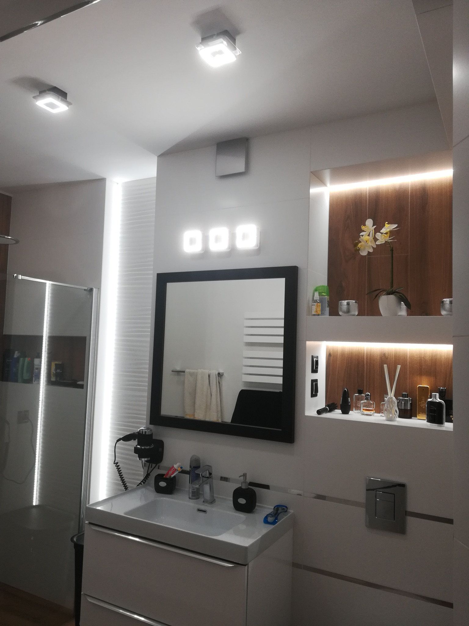 Nowoczesna łazienka z białymi płytkami, podświetlanym lustrem, umywalką z szafką, kabiną prysznicową z podświetleniem LED i dekoracyjną półką z drewnianym wykończeniem oraz kwiatami i perfumami.