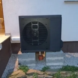 Zewnętrzna jednostka klimatyzacyjna Panasonic, zamontowana na betonowych blokach przed ścianą budynku, z widoczną rurką odprowadzającą skropliny i kamiennym obrzeżem rabaty.