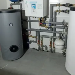 Kompleksowa instalacja grzewcza z szarymi rurami izolowanymi, zbiornikiem buforowym w szarej okładzinie i białym kotłem Aquarea Smart Cloud, zamontowana w kotłowni z szarą podłogą.