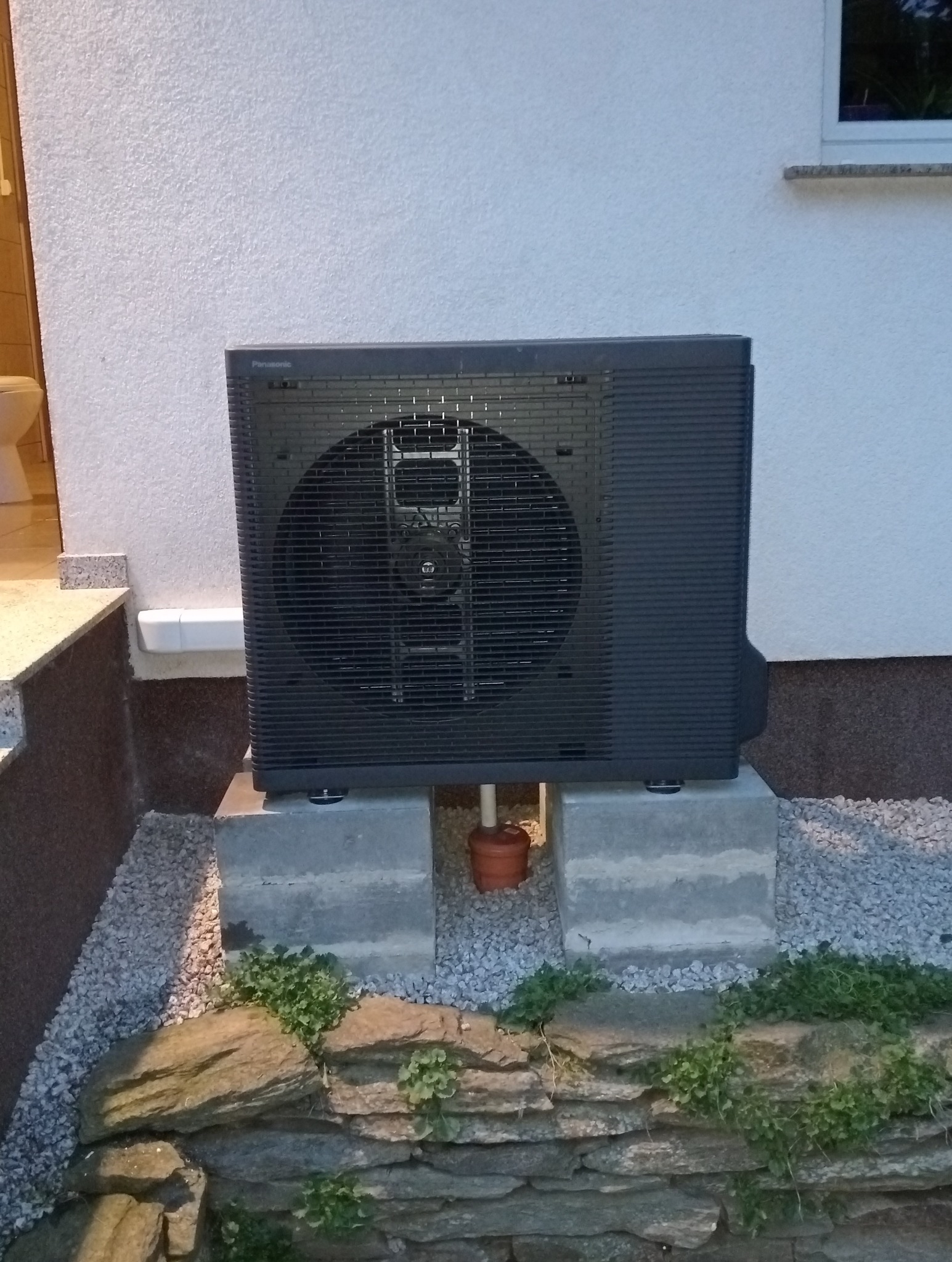 Zewnętrzna jednostka klimatyzacyjna Panasonic, zamontowana na betonowych blokach przed ścianą budynku, z widoczną rurką odprowadzającą skropliny i kamiennym obrzeżem rabaty.