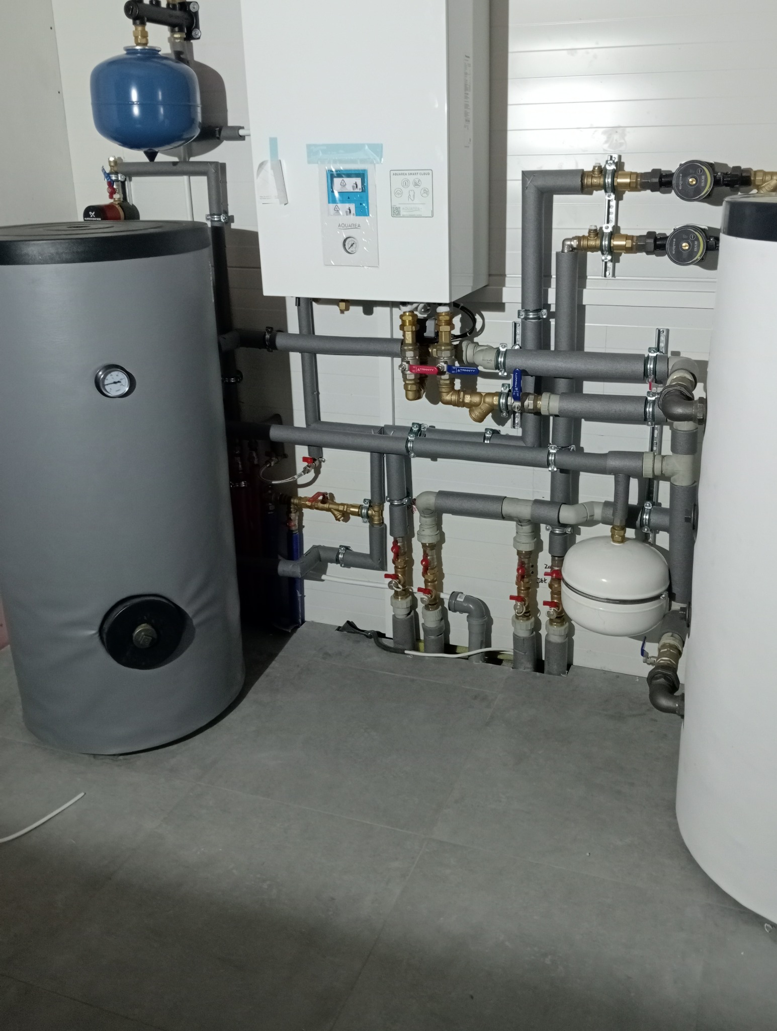 Kompleksowa instalacja grzewcza z szarymi rurami izolowanymi, zbiornikiem buforowym w szarej okładzinie i białym kotłem Aquarea Smart Cloud, zamontowana w kotłowni z szarą podłogą.