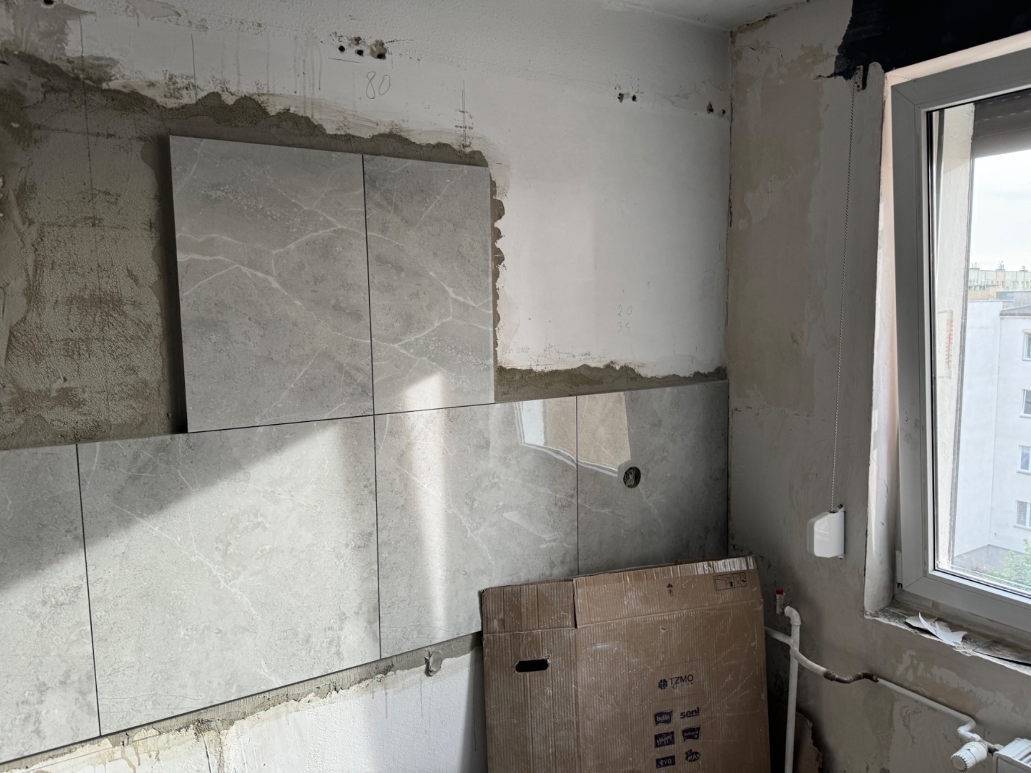 Układanie szarych płytek imitujących marmur na ścianie w trakcie remontu, widoczne ślady zaprawy i niedokończone fugi, kartonowe opakowanie po płytkach leży przy ścianie.