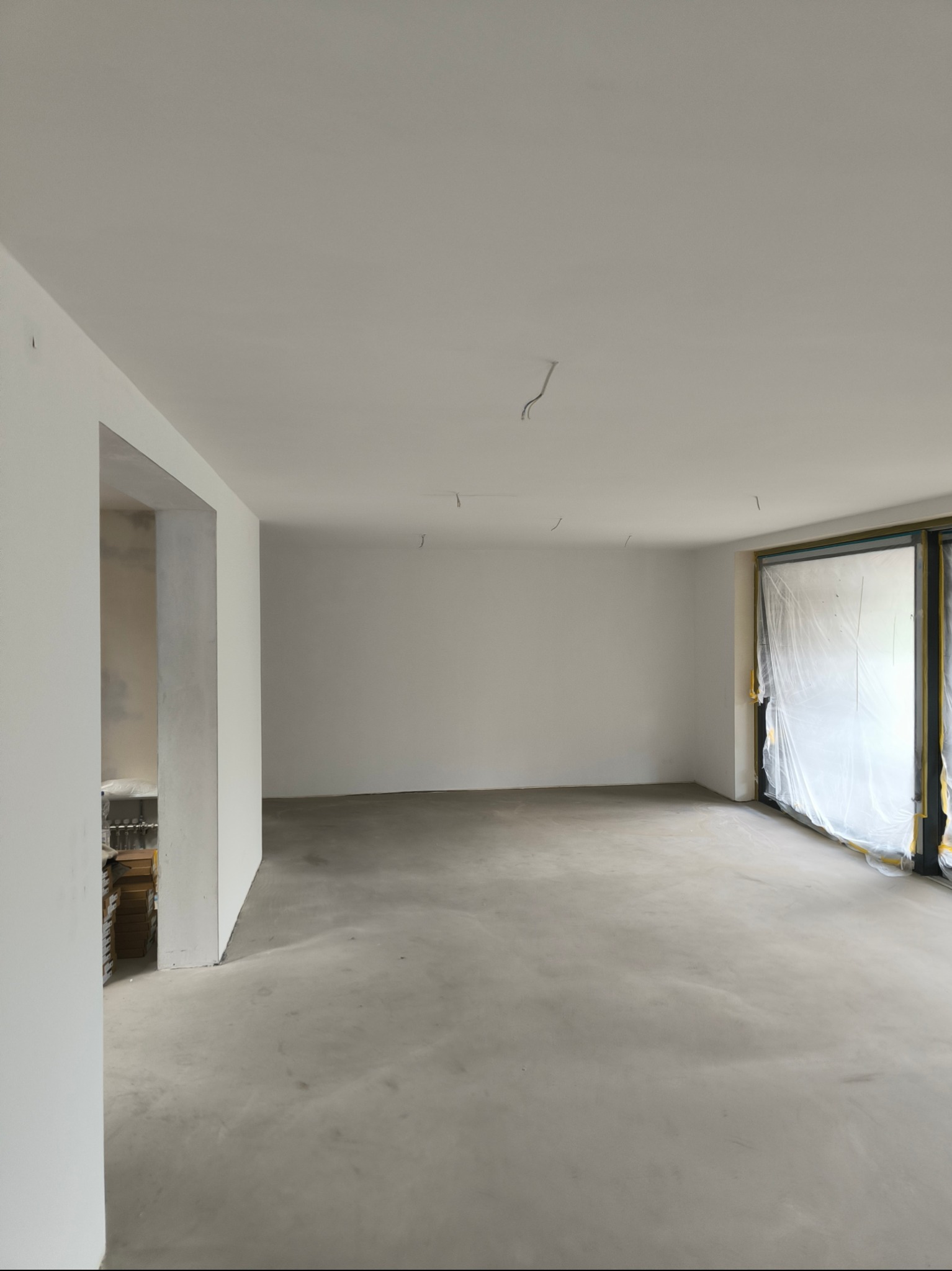 Pusty pokój z surowymi ścianami, przygotowany do prac wykończeniowych, okno zabezpieczone folią, widoczne wystające kable z sufitu, na podłodze wylewka.