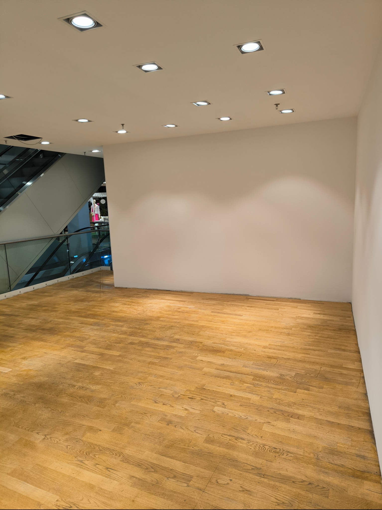 Pusta, jasna przestrzeń biurowa z parkietem w odcieniach brązu, białymi ścianami i sufitowymi lampami punktowymi, widok na schody ruchome w tle.
