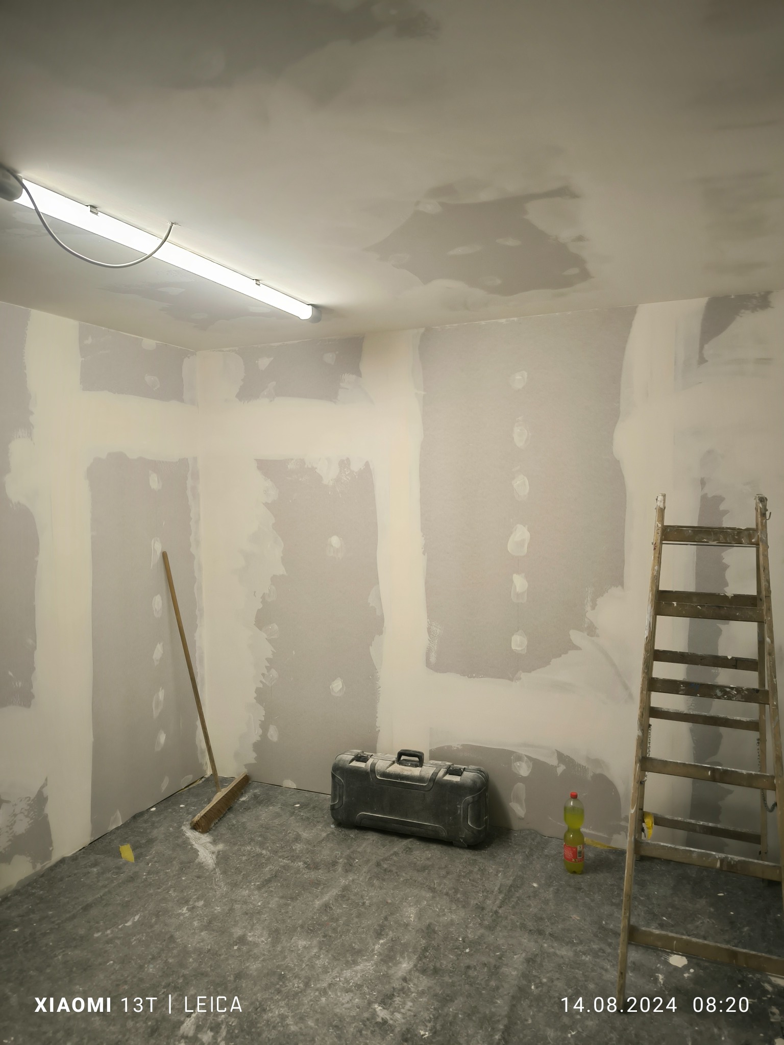 Pokój w trakcie szpachlowania ścian z płyt kartonowo-gipsowych, widoczna drabina, kufer z narzędziami i butelka z napojem na podłodze zabezpieczonej folią.