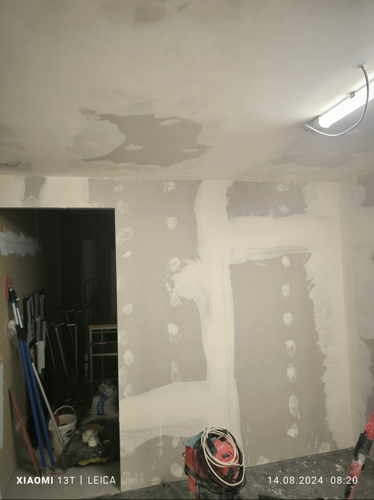 Pokój w trakcie szpachlowania ścian i sufitu, widoczne narzędzia i materiały budowlane, w tle otwarte przejście do innego pomieszczenia.