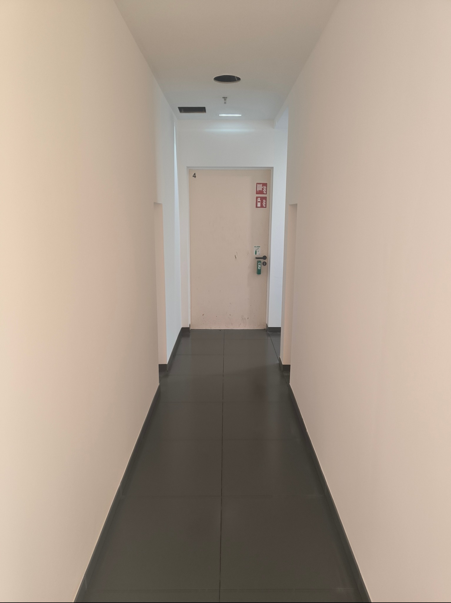 Korytarz biurowy z białymi ścianami, ciemną podłogą z płytek i drzwiami ewakuacyjnymi z numerem 4 i oznaczeniami przeciwpożarowymi.