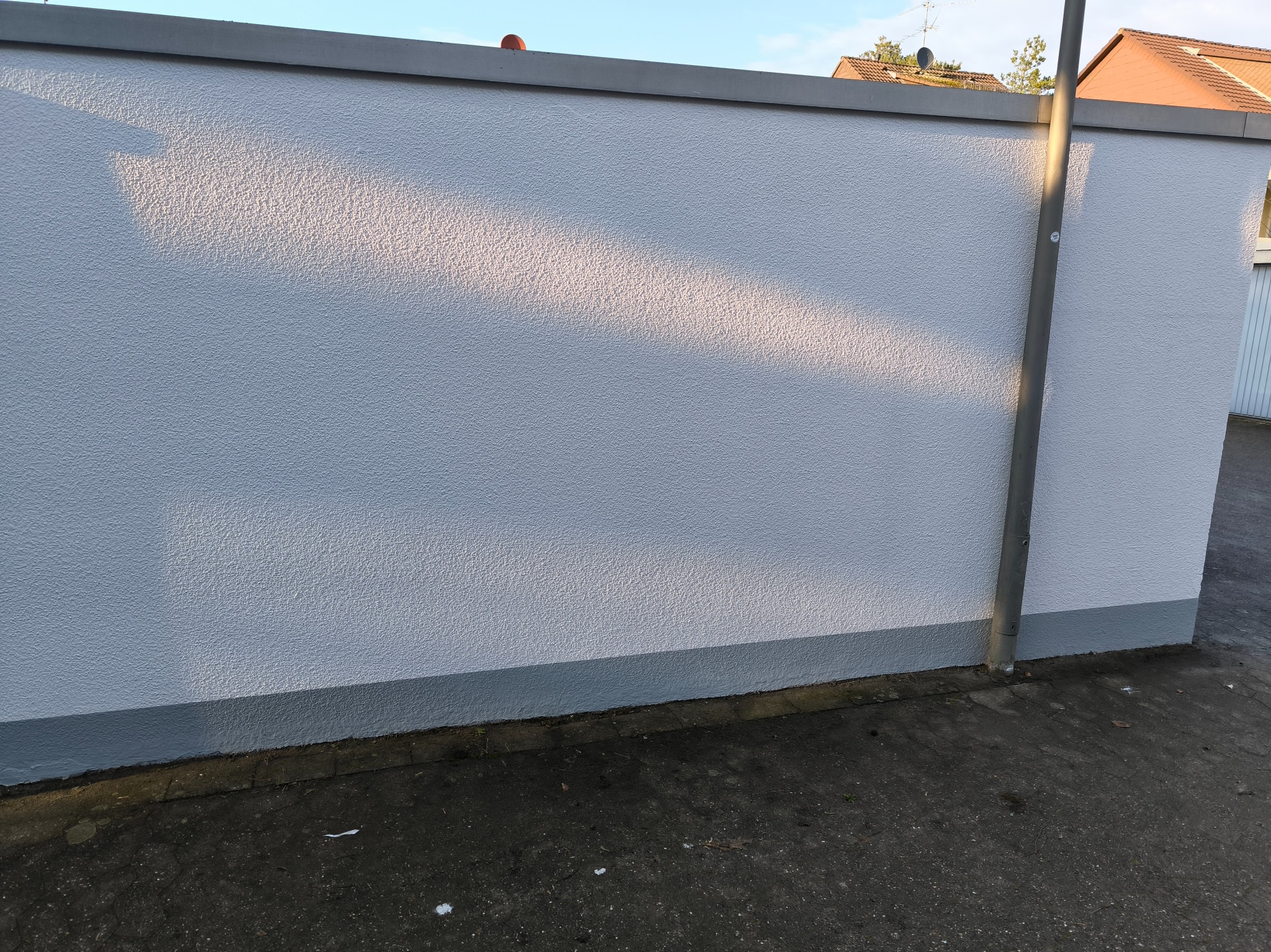 Świeżo otynkowana ściana zewnętrzna budynku z szarą listwą przypodłogową i górną, oświetlona popołudniowym słońcem, z widoczną rynną odprowadzającą wodę.