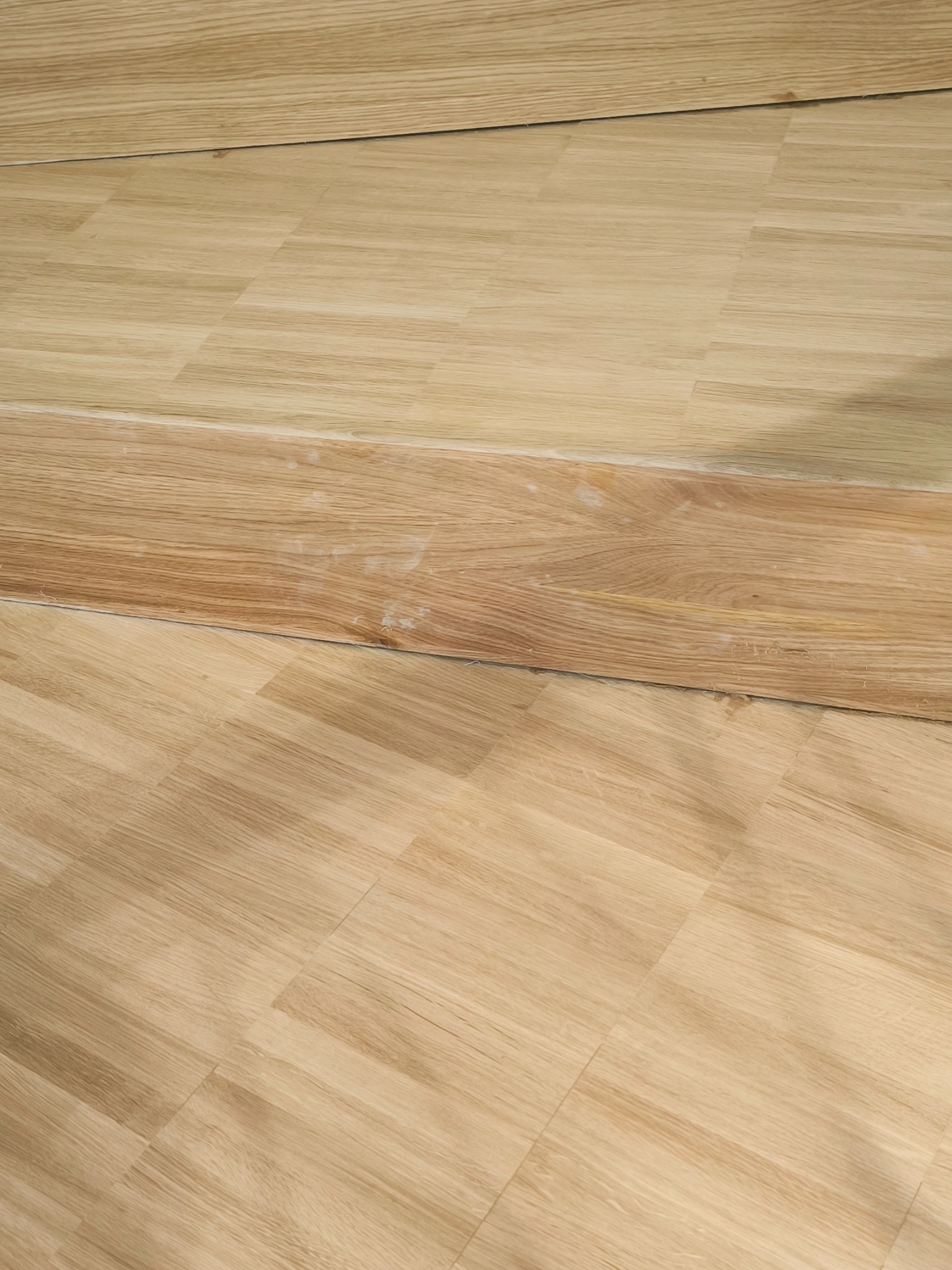 Fragment ułożonej, jasnej podłogi z parkietu, z widocznym fragmentem listwy przypodłogowej w innym odcieniu drewna, z delikatnymi śladami po kleju.